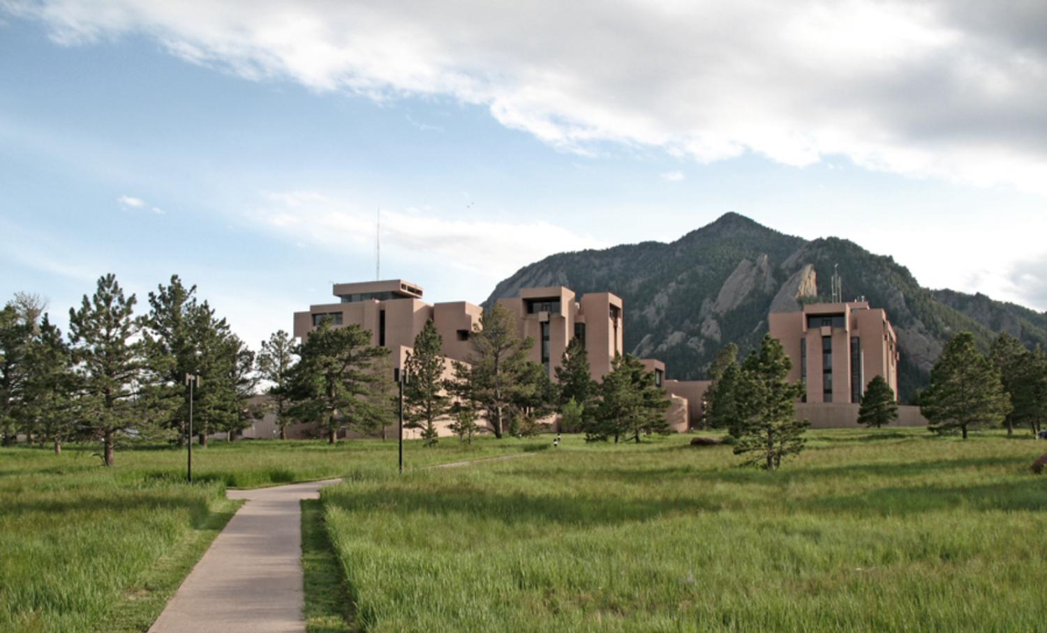 A path leads through green grass to the tall main buildings of NCAR, positioned in front of the Flatirons