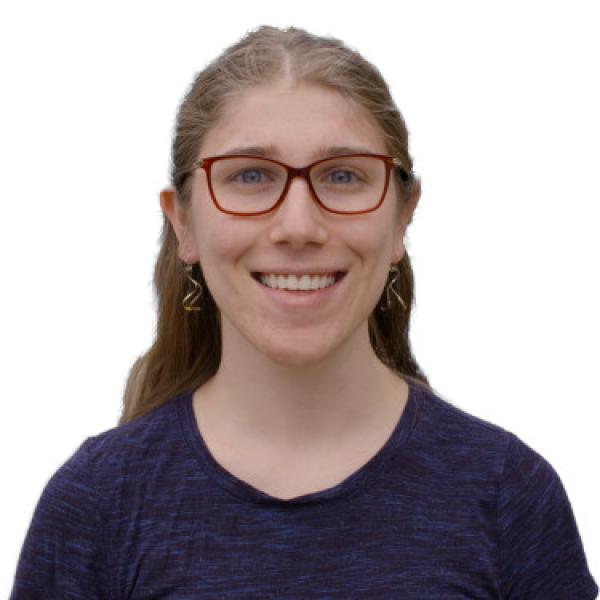 person with long brown hair hair, red squared eyeglasses, smiling, wearing a dark blue shirt and dangly earrings