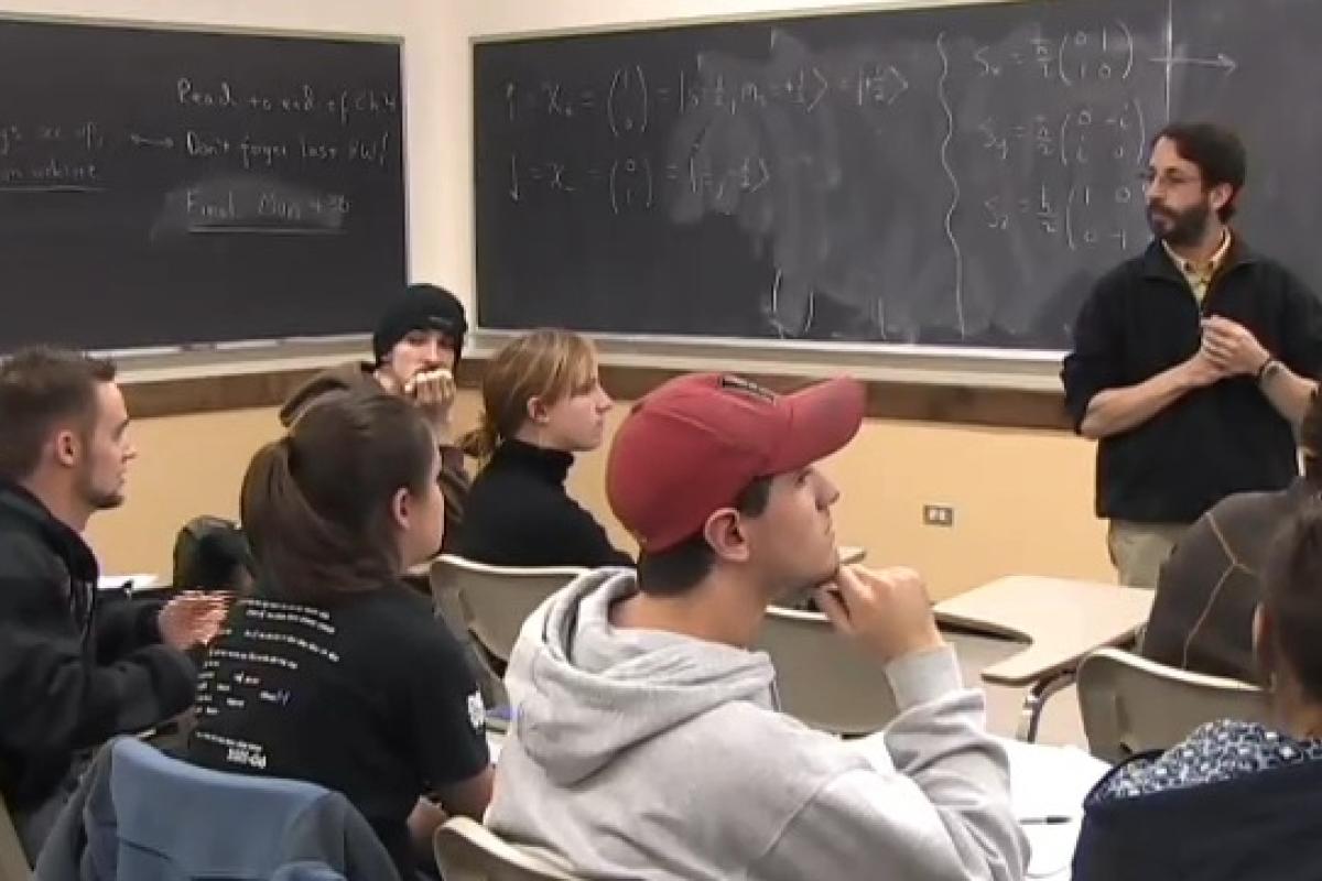 Students sitting at desks in a classroom listening to Professor Pollock who speaks in front of a chalkboard