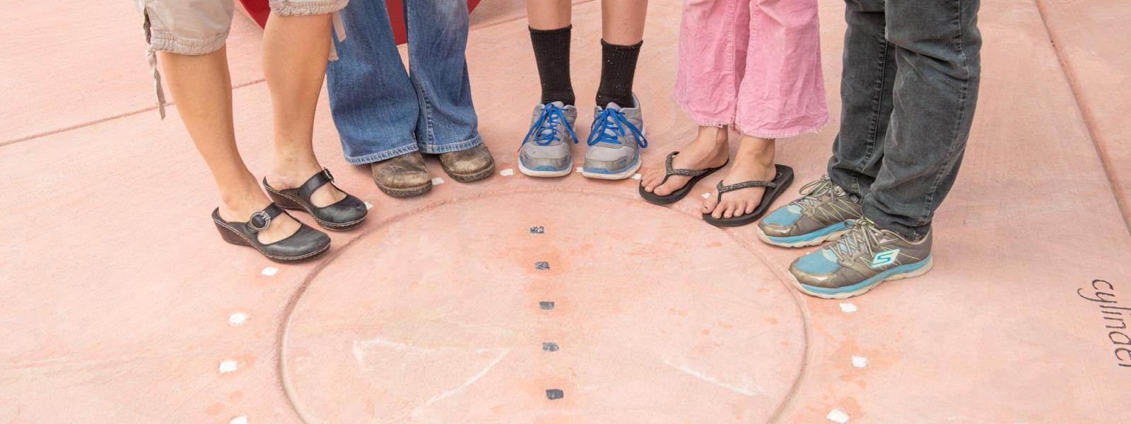 The shoes of several people can be seen, with them in the shoes, standing around a circle between some metallic dots.