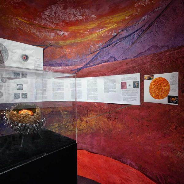 An orb-like object within a plexi vitrine on a black pedestal surrounded by a curved wall with bright red and orange coloring.