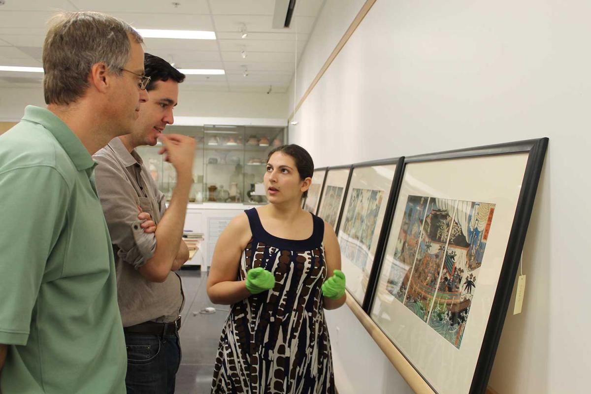 two male professors talk with a female students about framed prints on the wall in front of them