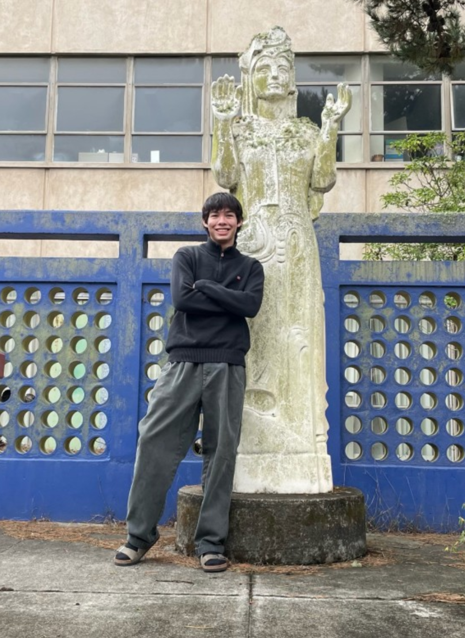 Tyler, who has pale skin and dark, straight hair. He is wearing dark sweatpants and a dark hoodie while smiling and standing next to a statue.