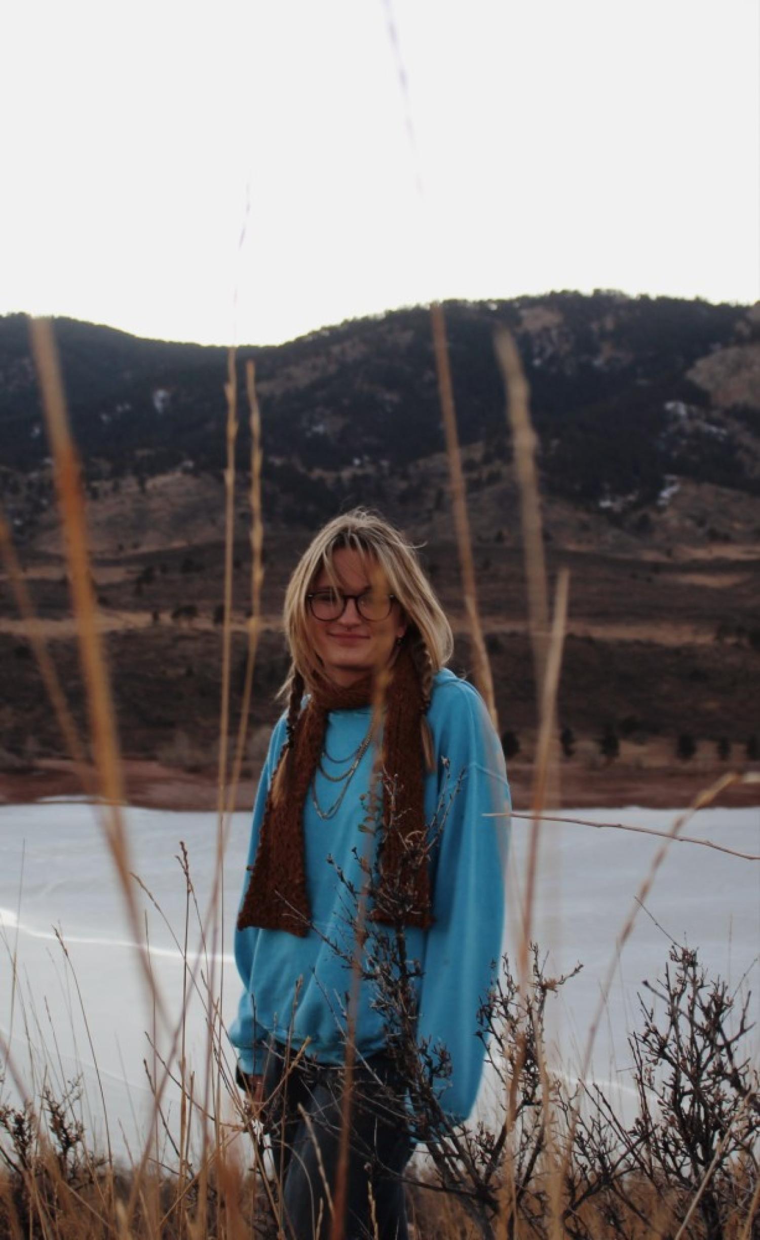 Anna, who is blonde with pale skin and is wearing glasses, a red scarf, and a blue sweater. She is standing outside by some ice in the mountains during an overcast day.