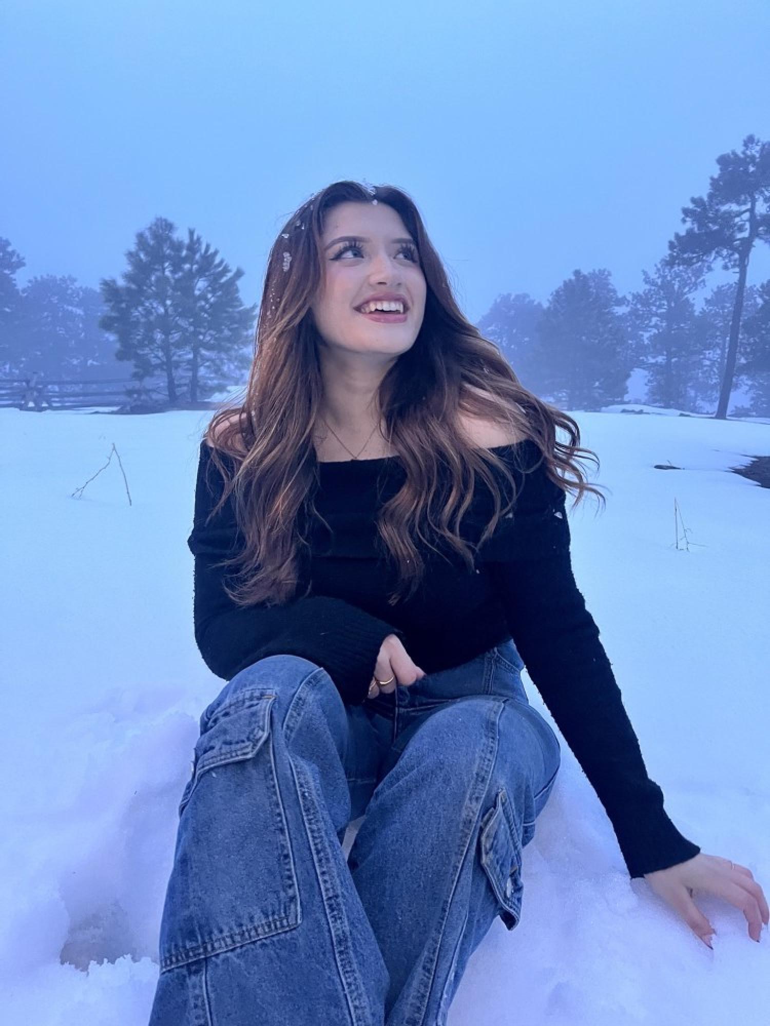 Nataly, who has fair skin and long, wavy brown hair. She is smiling, wearing a black sweater and jeans, and is sitting outside in the snow.