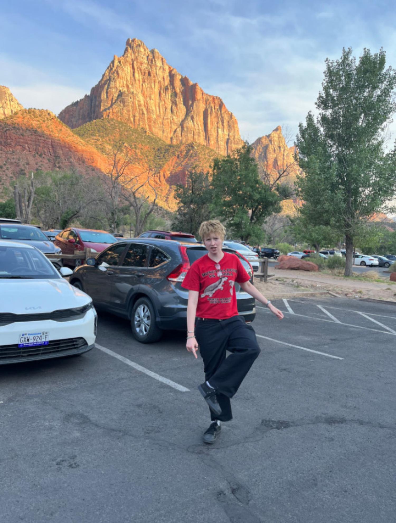 Quinn, standing outside in a parking lot with mountains in the background. He is wearing a red shirt and black pants and is pointing at his shoe. His hair is short and blonde.