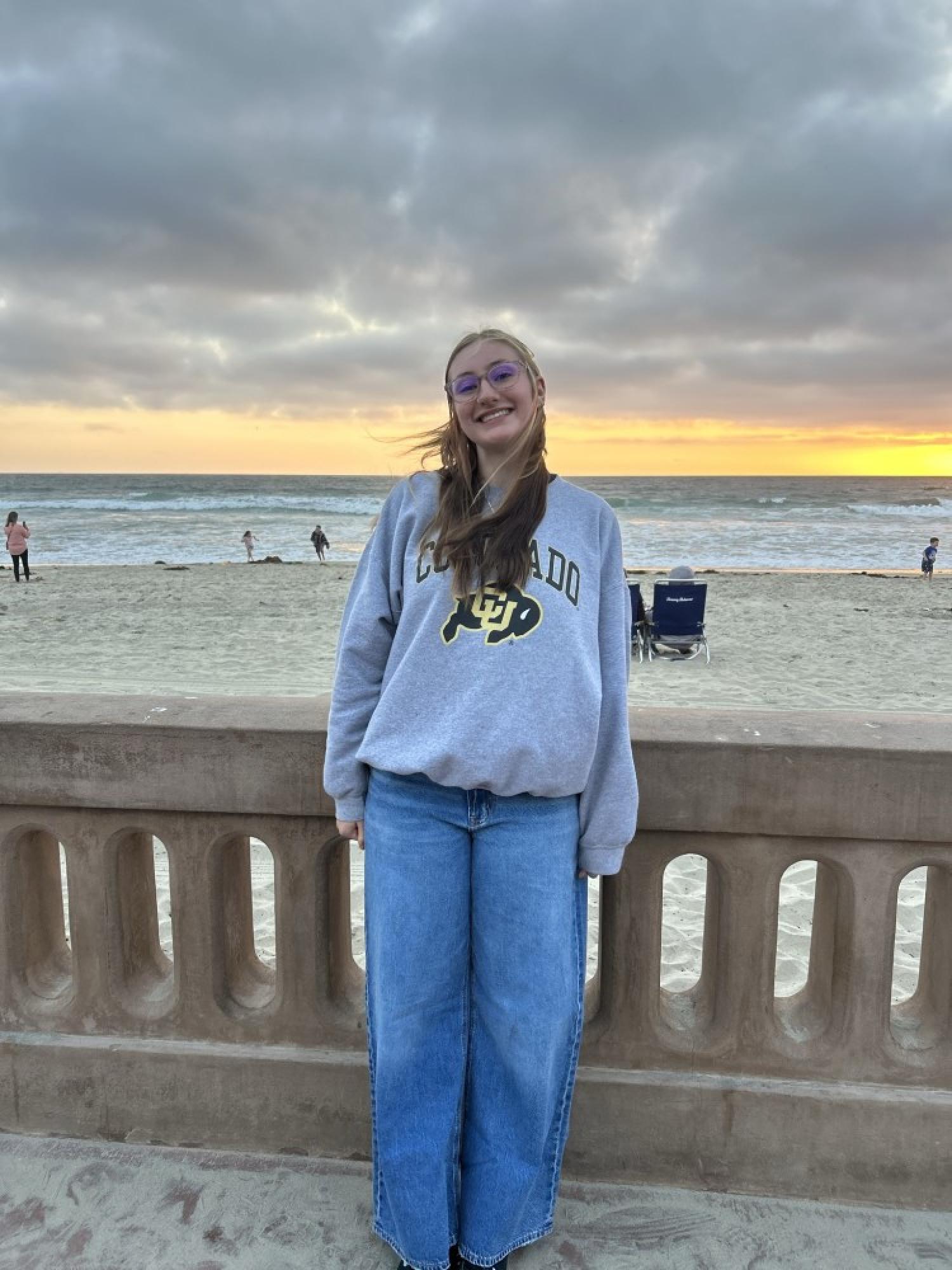 Rue-Water Brown, who is wearing a grey CU Boulder hoodie and jeans while standing outside at a beach at dusk. She is wearing glasses and smiling, and has pale skin and long, brown hair.