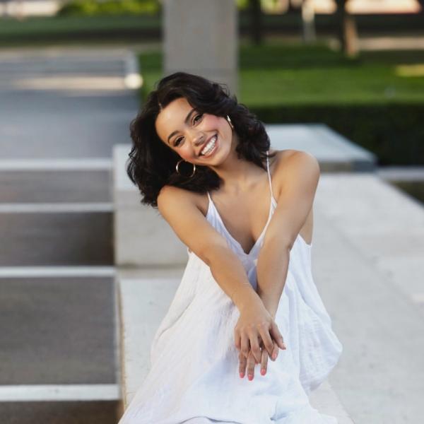 Ava, who has light skin and long, dark brown hair. She is wearing a white dress and is seated outdoors.