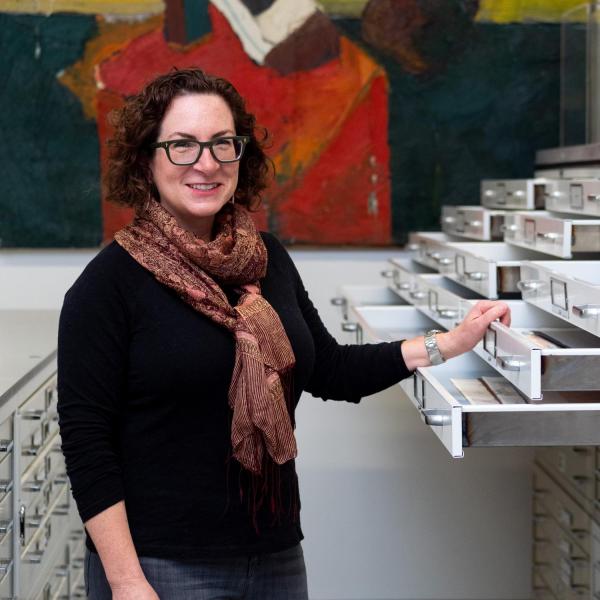 Hope Saska standing in the CU Art Museum vault. She has her hand rested on some open drawers in a flat filing cabinet and is wearing a dark sweater and a scarf.