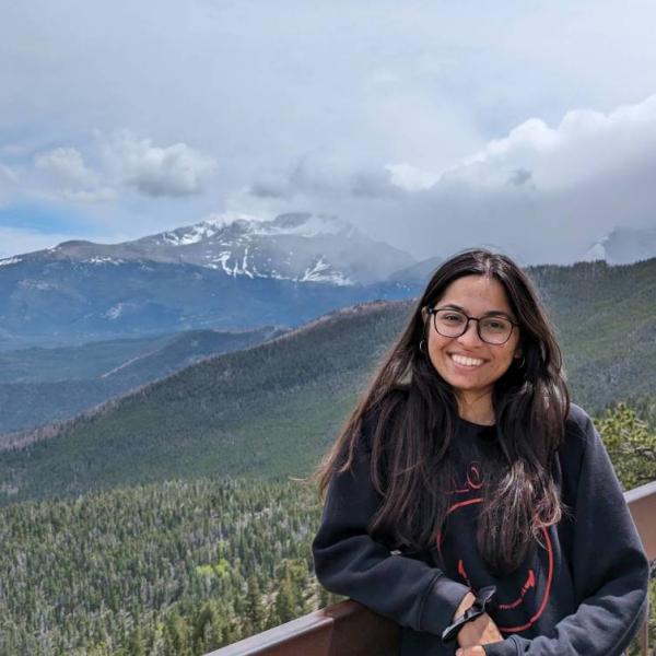 Nishee, a person with long dark brown hair, glasses, and light brown skin. She is wearing a dark, long sleeve shirt and jeans and is outdoors with mountains in the background.