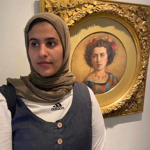 Rawan, standing next to an ornately framed painting depicting a woman with a flower crown. Rawan is wearing a beige hijab and a dark vest over a long sleeve white T-shirt.