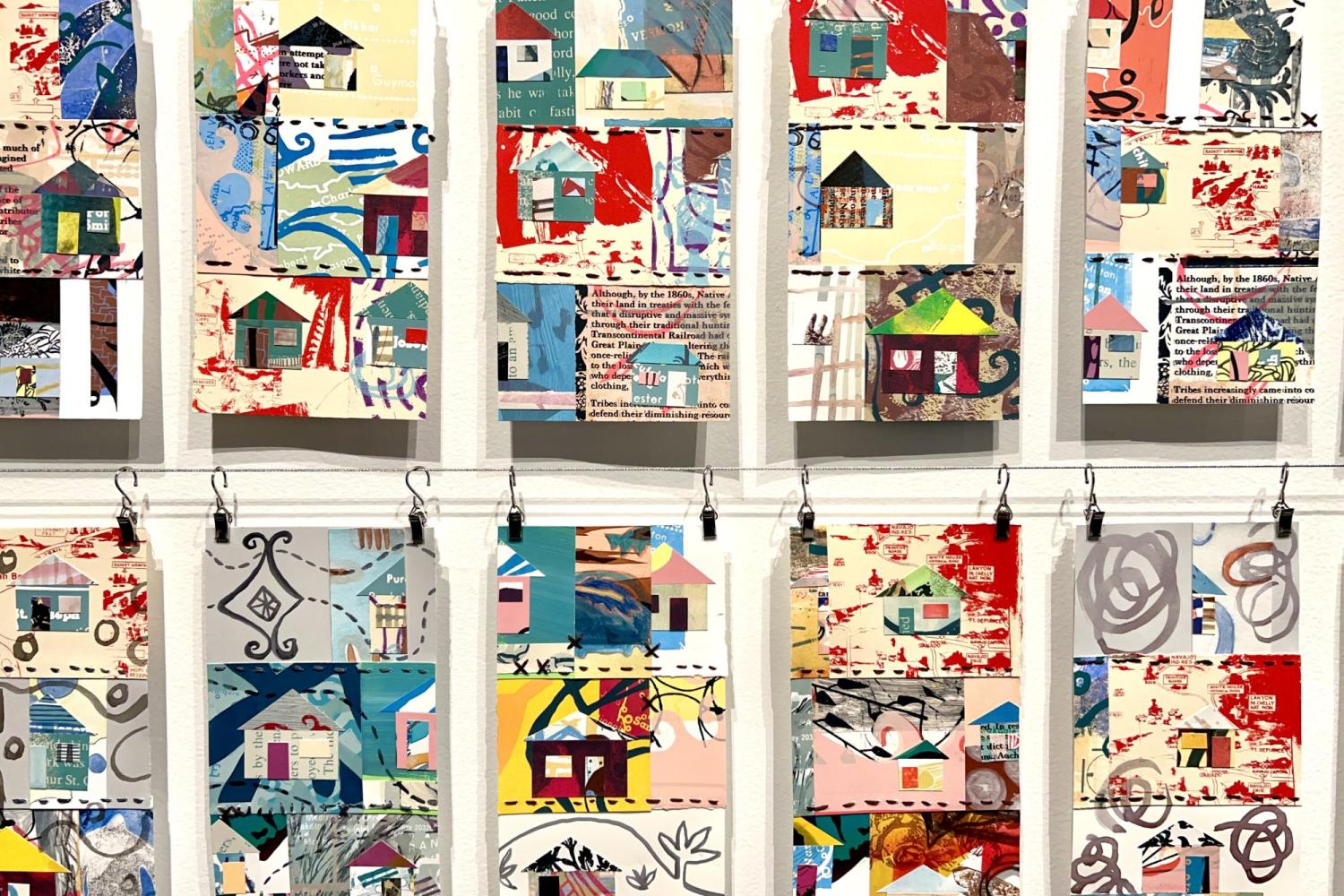 Many rectangular pages of collaged paper with colorful houses, other designs and text clipped to a metal line and hanging in front of a white wall.