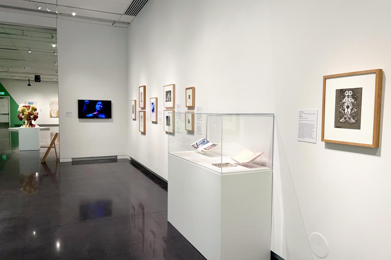 A photo of the inside of the CU Art Museum, featuring a wall with many framed artworks, a TV screen mounted on the wall, and a pedestal with artwork inside.