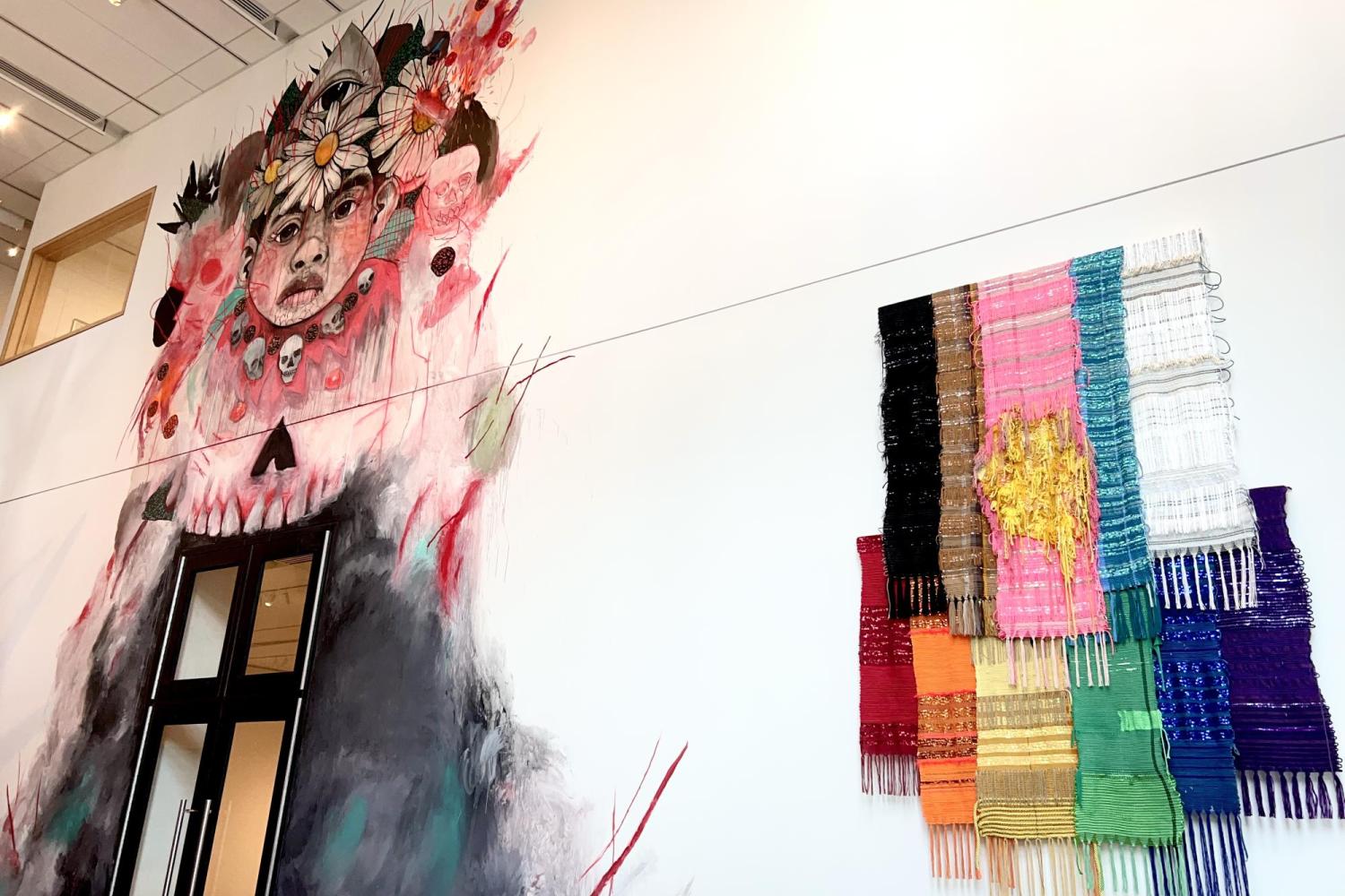 The large white lobby wall of the CU Art Museum, featuring a mural depicting a child wearing a necklace of skulls and a flower crown above the doors on the left, and a hanging rainbow flag made of knit fabrics, sequins, cords, and tassels to the right.