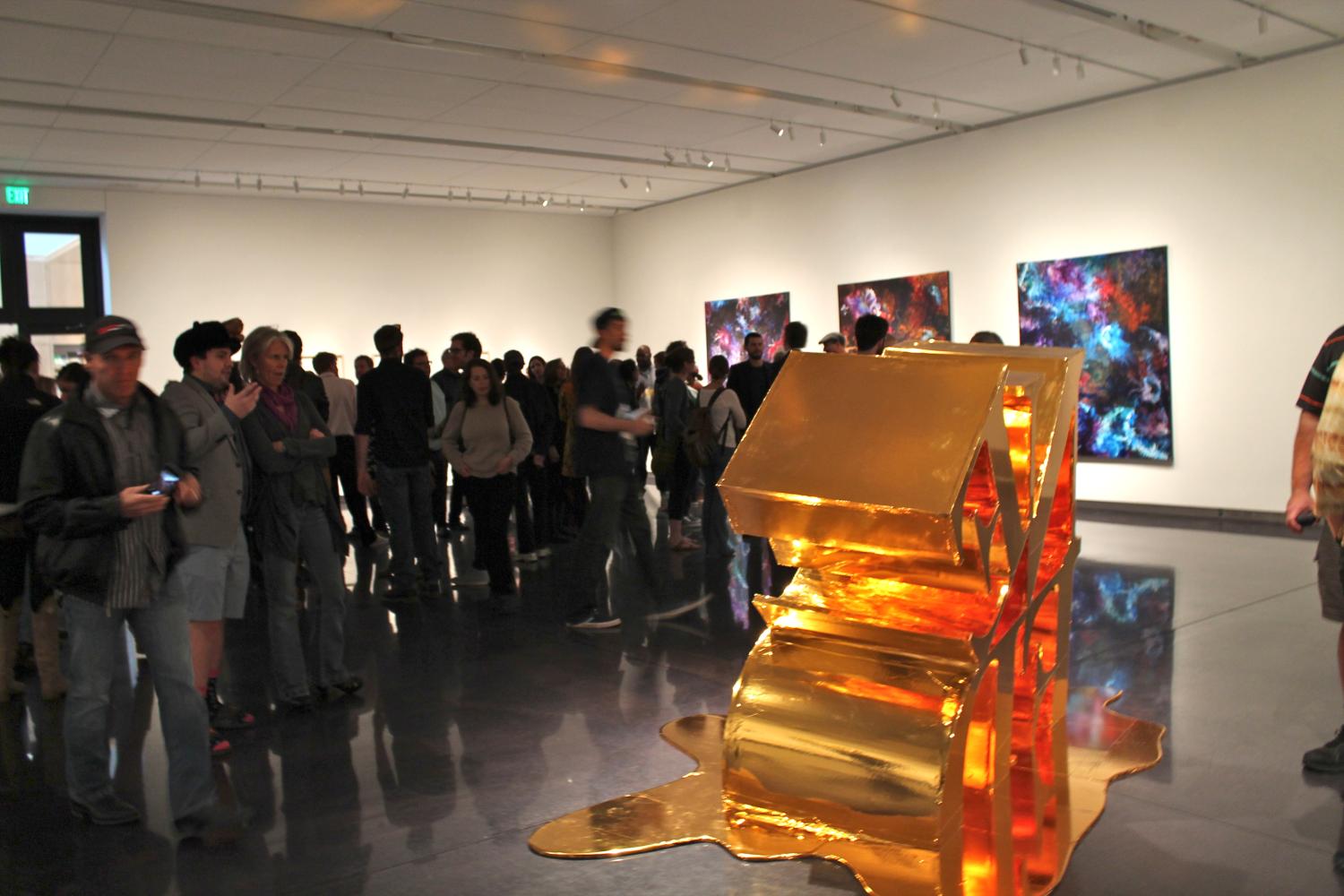 A crowd mingles in the gallery with a large gold sculpture in the foreground.