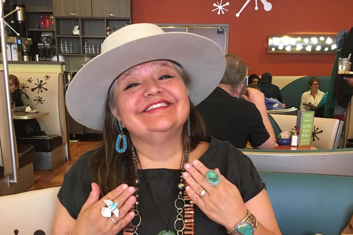 Melanie Yazzie, a Native American woman with dark hair with grey streaks. She is smiling and wearing colorful jewelry, including torquoise, and a white, brimmed hat.