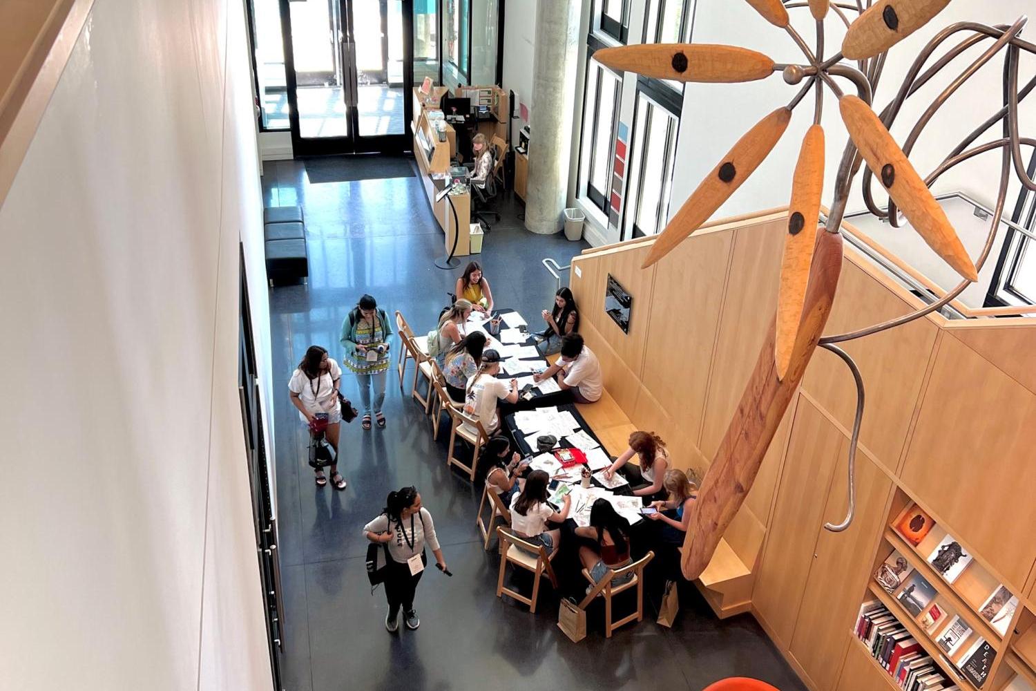 A view from above of the CU Art Museum lobby. There is a long table with a black tablecloth on it at which many people are seated and working on coloring pages together. In the foreground is a large metal and wood sculpture suspended from the ceiling.