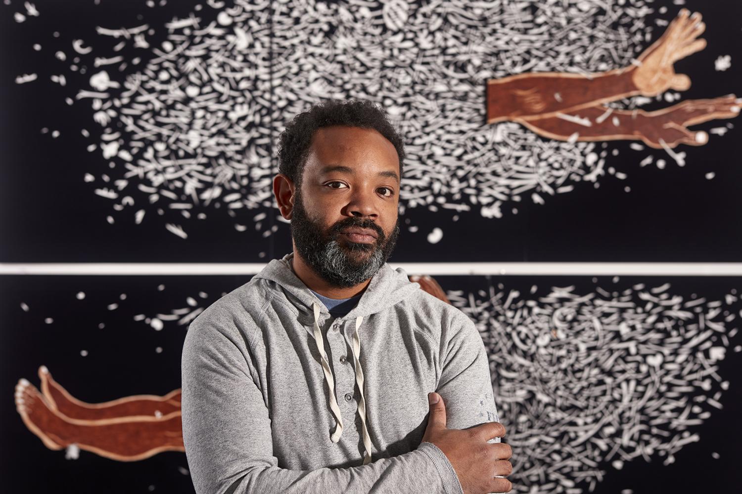 William Villalongo, a black man with short, dark hair and a greying beard, standing in front of some of his artwork featuring outstretched limbs amidst a dark background with white filigree cutouts.