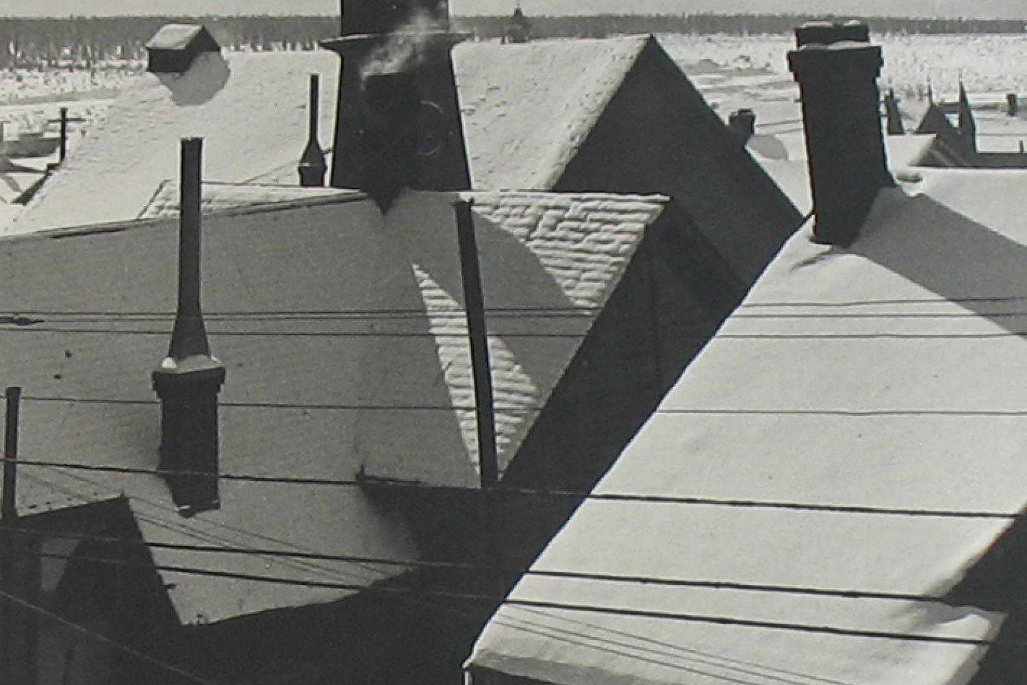 A black and white photograph of several snowy roofs. In the far distance are mountains.