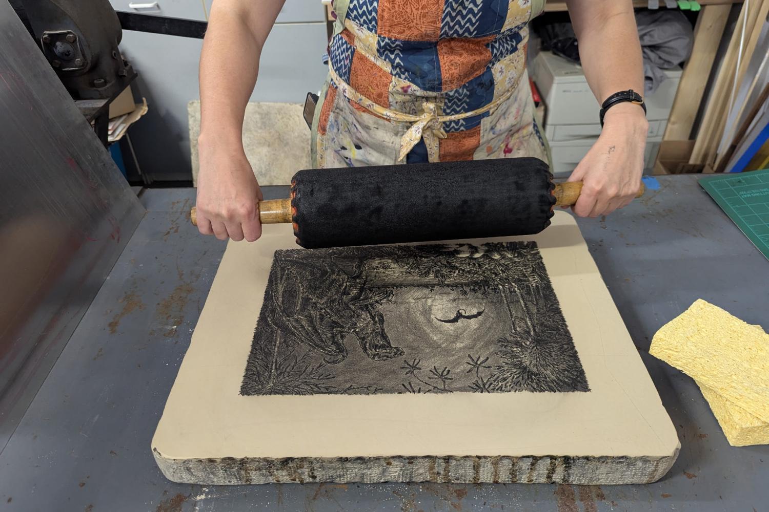 A woman using a roller to roll ink onto a lithography stone with a landscape drawn on it.