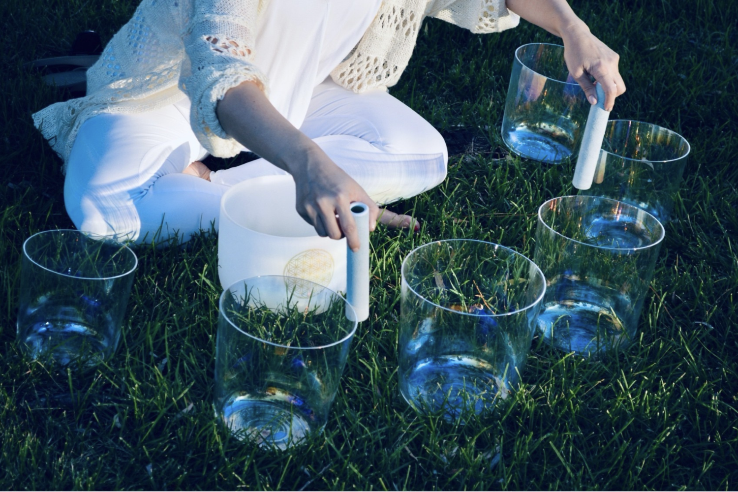 A person dressed in white sitting on the grass while touching several clear, quartz sound bowls.
