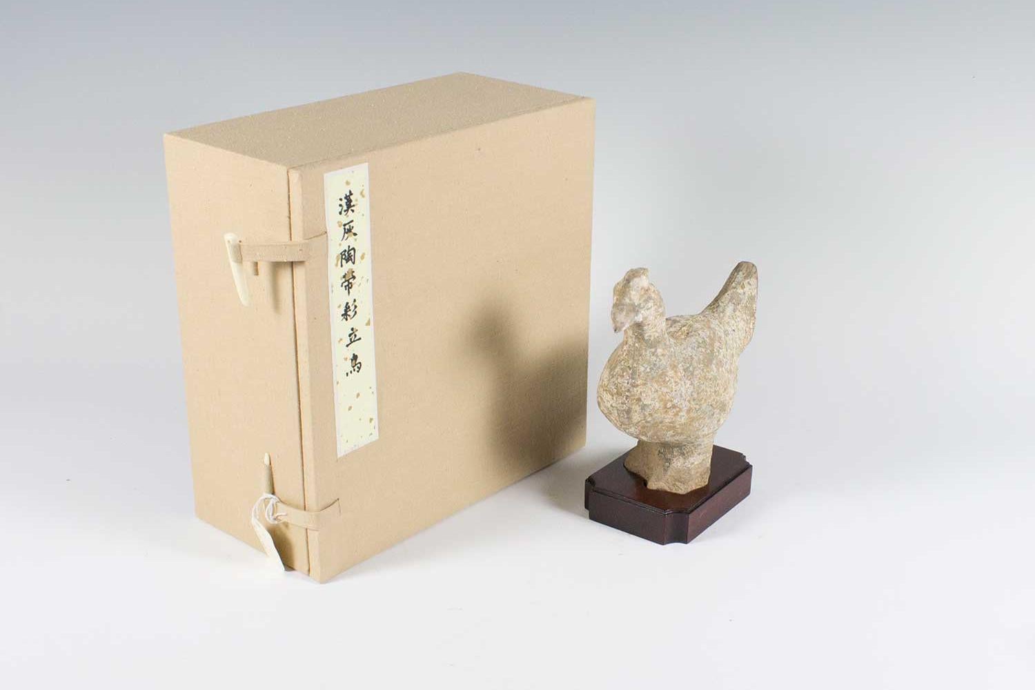 Clay figure of a small chicken standing on a wooden stand with tan box on left.