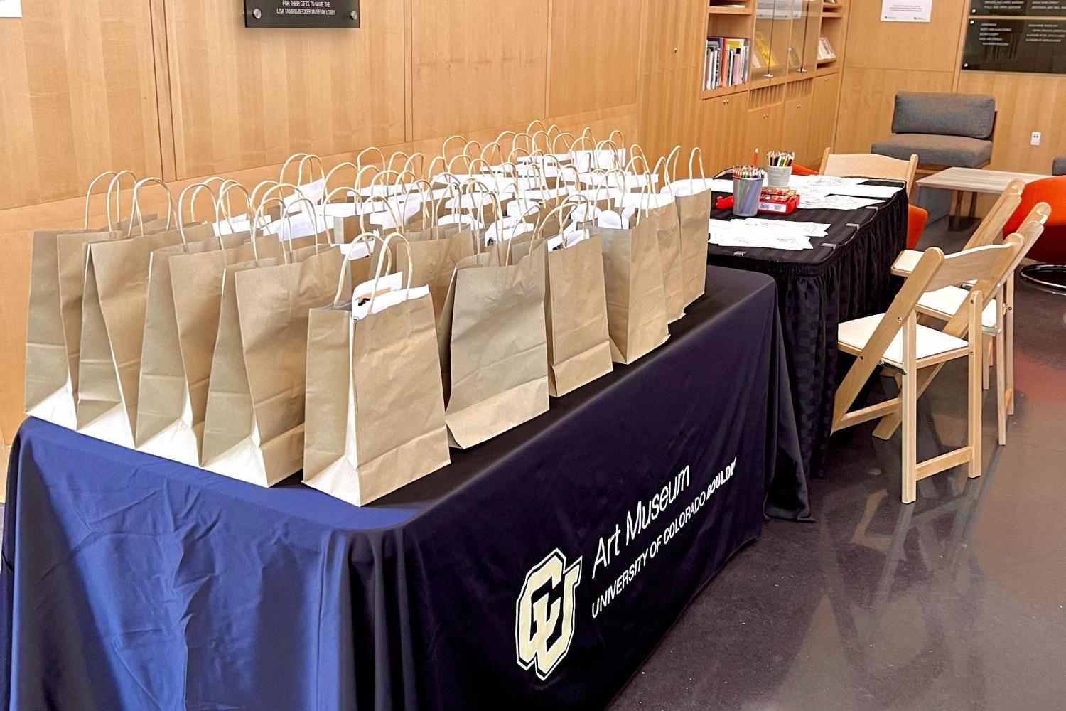 Many brown paper bags lined up on a table in the CU Art Museum lobby. They contain art supplies!