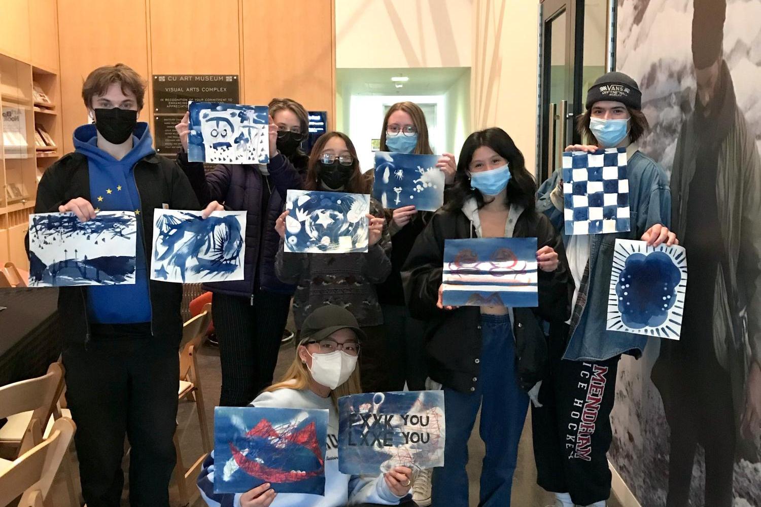 Many people wearing masks and standing in a group while holding up cyanotypes they created. They are in the CU Art Museum Lobby together.