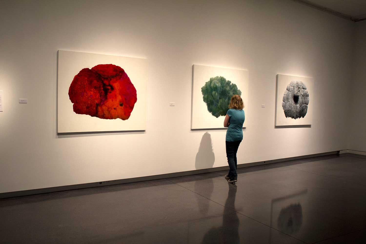 a woman stands in front of three large drawings hanging on the wall, one is red, one is green, on is grey