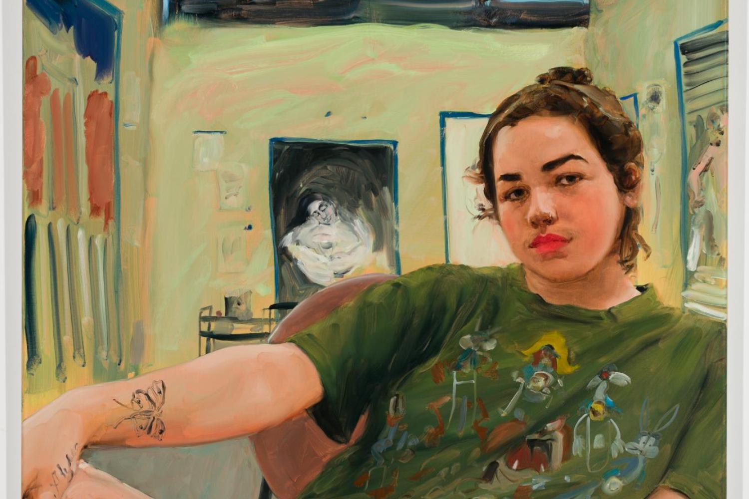 A painting of a person reclining with their leg up and bent. They have light skin and brown hair pulled into a messy bun. They are wearing a dark green graphic T-shirt and black shorts. They have many black tattoos on their arms and legs and are seated in a greenish room with paintings hanging from the walls, their studio.