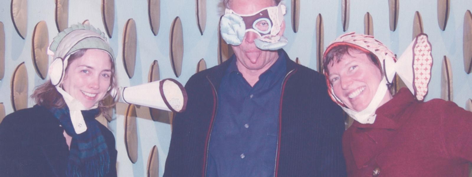 Three people standing next to each other wearing whimsical hats and masks. They are making silly faces. They are, from left to right: Kim Dickey, Scott Chamberlain, and Jeanne Quinn.