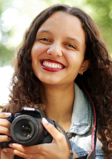 St. Clair smiling and holding a camera. She is black and has long curly hair.