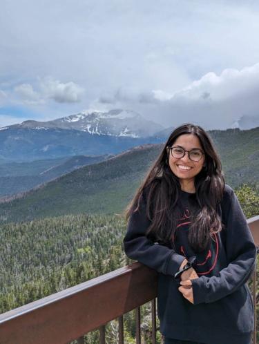 Nishee, a person with long dark brown hair, glasses, and light brown skin. She is wearing a dark, long sleeve shirt and jeans and is outdoors with mountains in the background.