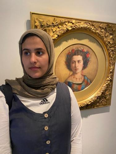 Rawan, standing next to an ornately framed painting depicting a woman with a flower crown. Rawan is wearing a beige hijab and a dark vest over a long sleeve white T-shirt.