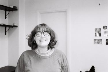 A black and white photo of Riley. Her eyes are closed and she is smiling. She has short hair with bangs, and is standing indoors with a door and some shelves behind her.