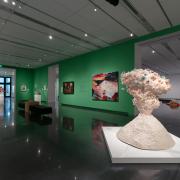 An installation photo of the inside of the CU Art Museum's galleries, featuring a bright green wall and many colorful nature-themed artworks. Paintings, prints, and other works are framed and hanging on the walls, while scultures rest on pedestals around the room. Two black benches and a cube-like wooden table are in the center of the room.