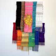 A rainbow textile featuring tassels, sequins, and cords hanging from a while wall. The top of the textile features stripes (from left to right) of black, brown, bright pink, light blue, and white. On the bottom (from left to right) are stripes of red, orange, green, blue, and purple. In the middle of the top layer is a large yellow circle with lots of loop cords and string.