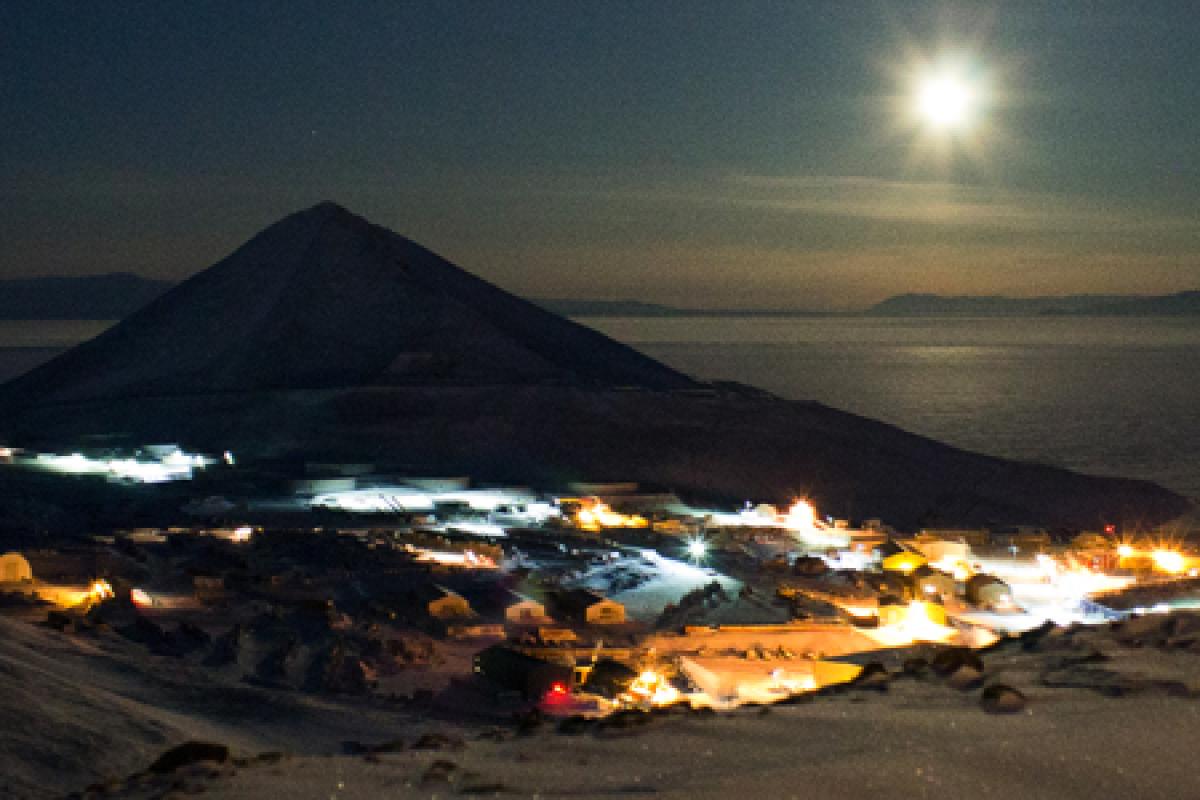 McMurdo Station at the base of a mountain by the bay with moon in the distance.