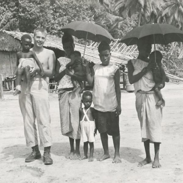 Bud holding baby next to three women on beach, along with two more babies and a toddler. The women are holding sun umbrellas.