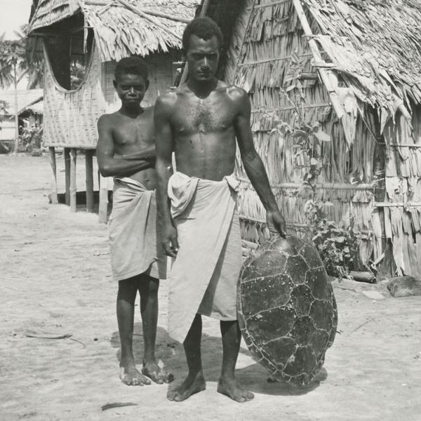 Male youth and man holding large turtle shell on beach.