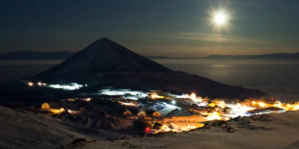 McMurdo Station at the base of a mountain by the bay with moon in the distance.