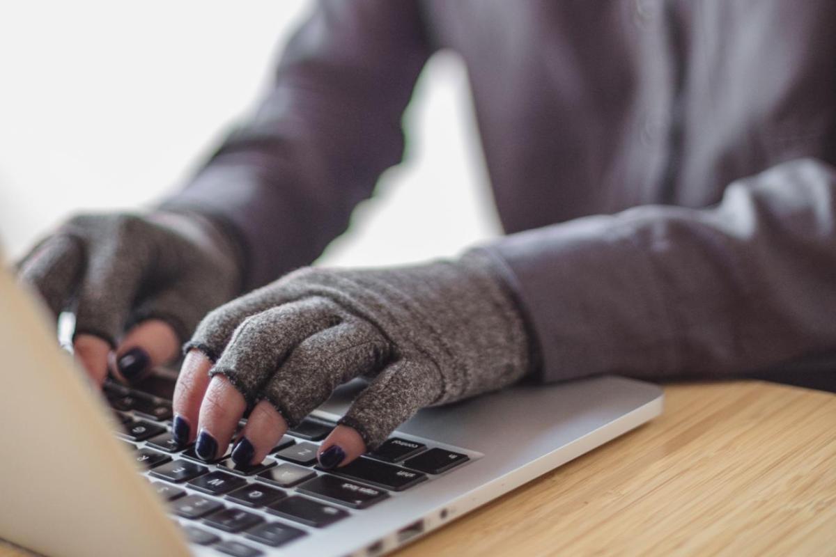 A disabled Asian genderfluid person types on a laptop while wearing compression gloves. The hands and keyboard are the focal point.