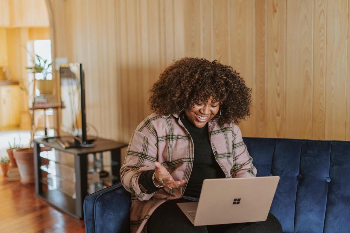 Person with curly brown hair sitting on couch smiling in front of laptop