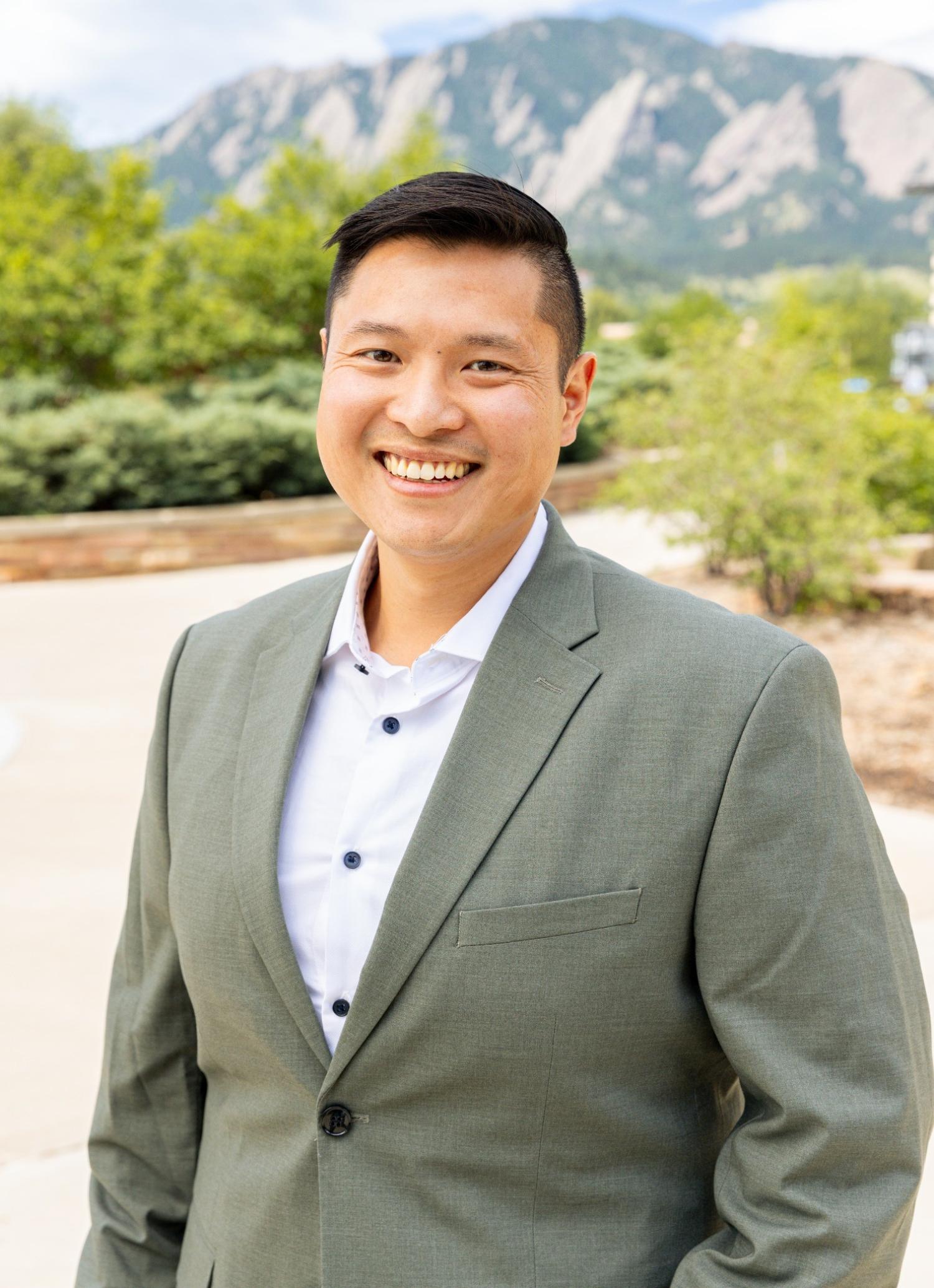 Headshot of Stanley Ly in a suit jacket posed in front of Flatiron Mountains.