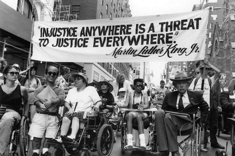 A group of activists with varying disabilities, racial identities, genders, and age demographics holding a sign that reads ‘Injustice anywhere is a threat to justice everywhere. Martin Luther King Jr.’