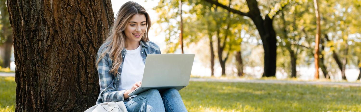 Photo of a student on a laptop outside under a tree on a sunny day.