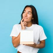 Photo of a student standing in front of a blue background with a pondering look as they hold up a calendar.