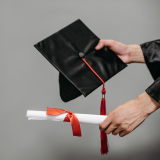 one hand holding a graduation cap with a red tassel and the other hand holding a rolled paper with a red ribbon