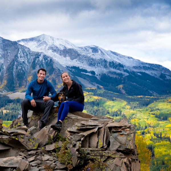 Sarah with her husband and dog on a rocky point with snow-covered mountains and fall leaves in the background