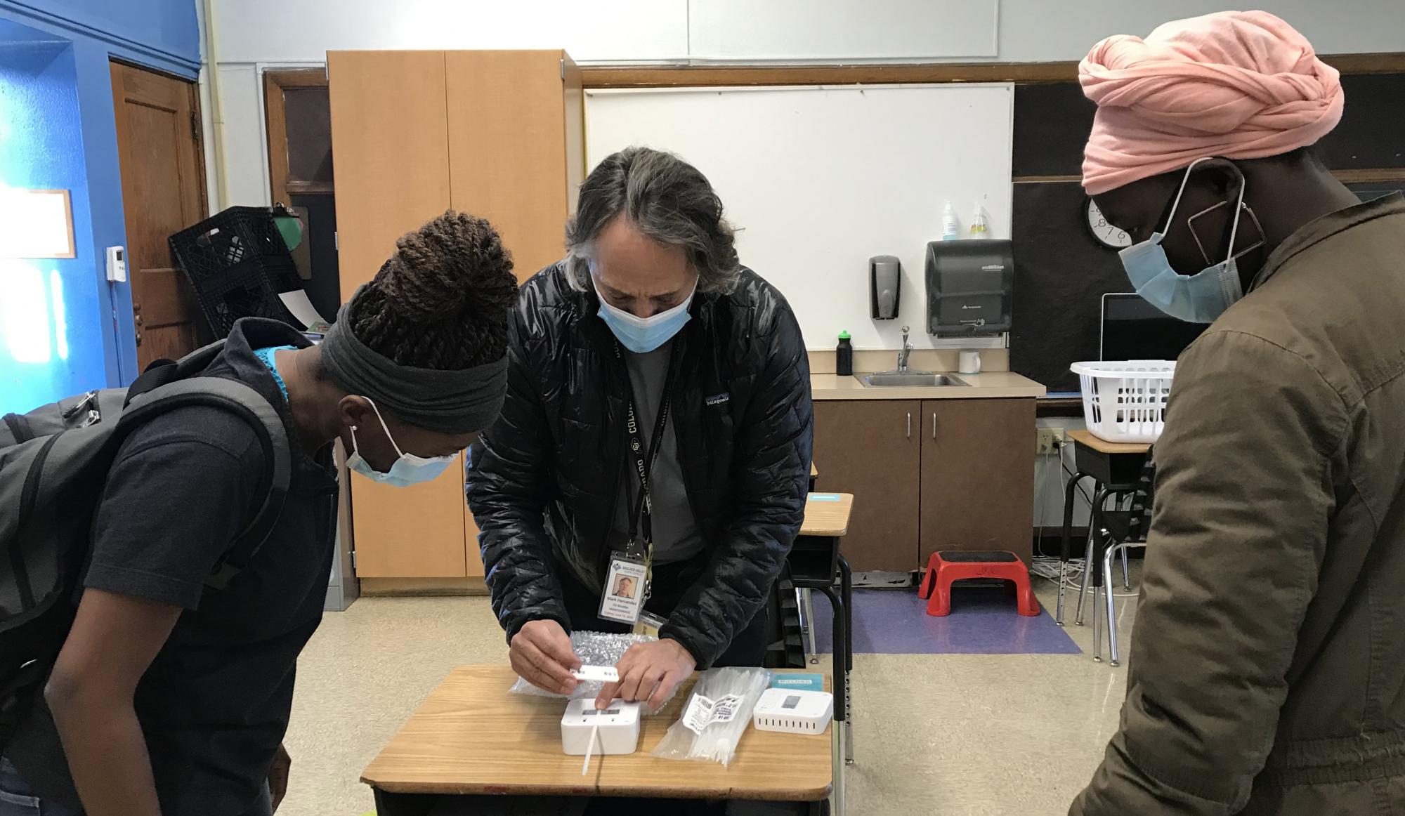 Processor Mark Hernandez demonstrates the installation process for the classroom air quality remote sensors to CU Boulder student volunteer technicians Christiane Nitcheu and Sylvia Akol.